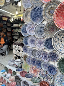 A market stall displays a wide array of brightly colored, intricately patterned ceramic plates, bowls, and pottery. The designs are primarily geometric and floral, featuring dominant hues of blue, red, green, and white. To the left, a person is seated, wearing a hoodie and a cap, with bags and other pottery items in the background.