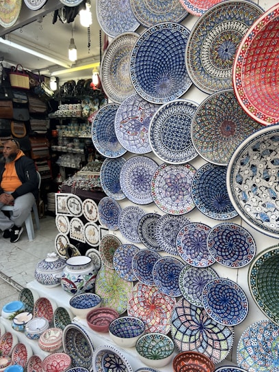 A market stall displays a wide array of brightly colored, intricately patterned ceramic plates, bowls, and pottery. The designs are primarily geometric and floral, featuring dominant hues of blue, red, green, and white. To the left, a person is seated, wearing a hoodie and a cap, with bags and other pottery items in the background.