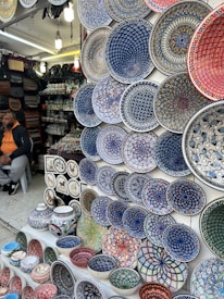 A market stall displays a wide array of brightly colored, intricately patterned ceramic plates, bowls, and pottery. The designs are primarily geometric and floral, featuring dominant hues of blue, red, green, and white. To the left, a person is seated, wearing a hoodie and a cap, with bags and other pottery items in the background.