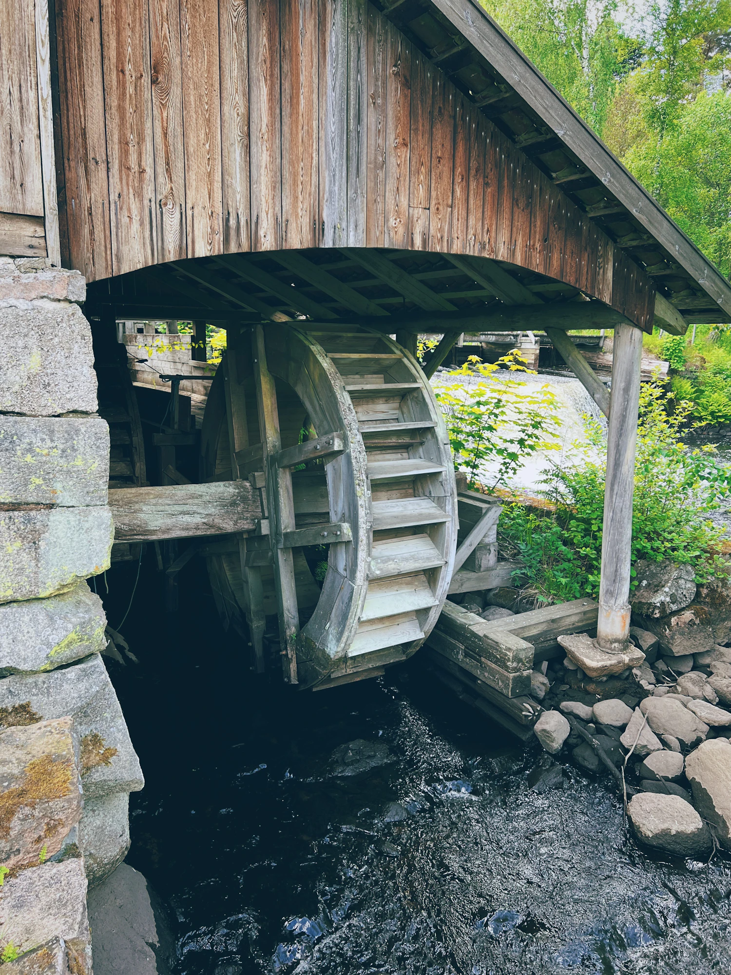 A timber mill building standing over dark water, its wheel shaded under the eaves