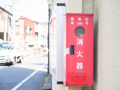 A red fire hose box mounted on a beige wall with Japanese text. The background features an urban street scene with buildings and a white truck parked on the side of the road under a clear sky.