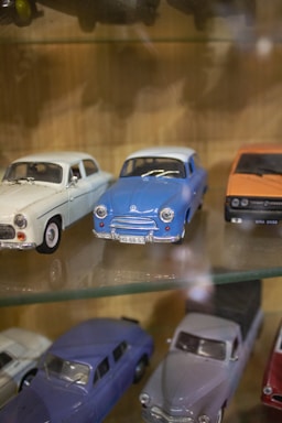 A collection of vintage toy cars is displayed on a glass shelf. There are white, blue, and orange cars in various designs, showcasing the classic automotive styles. The detail on the miniature models highlights the craftsmanship involved in creating these toys.