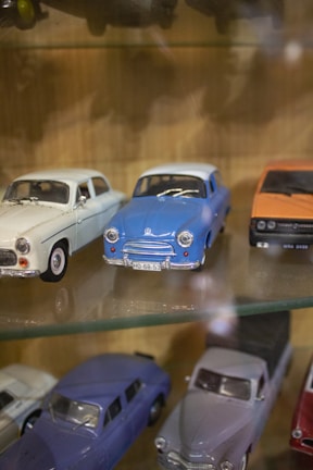 A collection of vintage toy cars is displayed on a glass shelf. There are white, blue, and orange cars in various designs, showcasing the classic automotive styles. The detail on the miniature models highlights the craftsmanship involved in creating these toys.