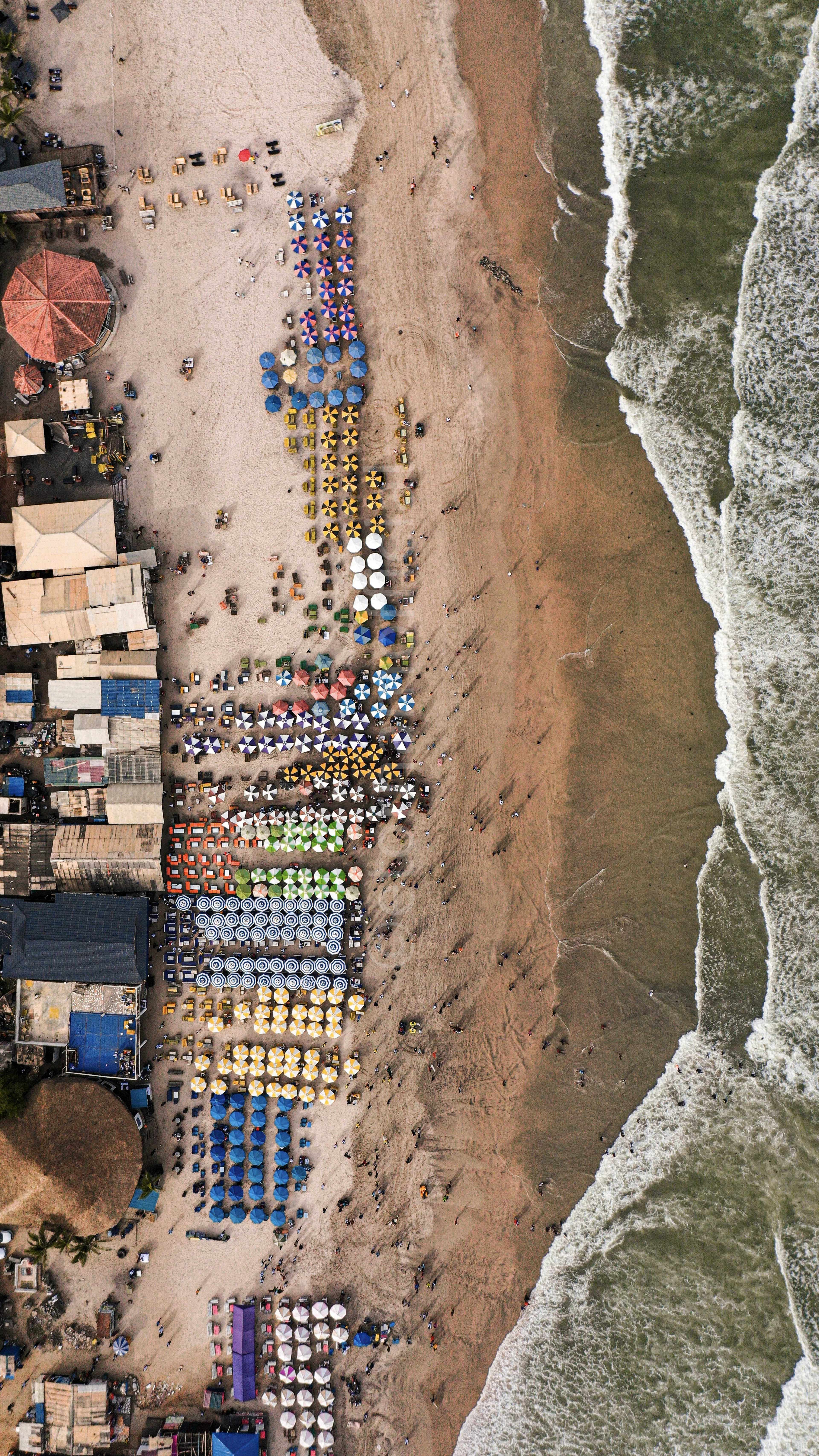 Aerial view of colorful beach umbrellas lined in rows along the sand beside breaking ocean waves.