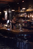 A dimly lit bar scene with two bartenders preparing drinks behind a wooden counter. The bar is stocked with various bottles on shelves, and the lighting creates a cozy atmosphere. Several hanging pendant lights illuminate the area, and there are bar stools lined up in front of the counter.