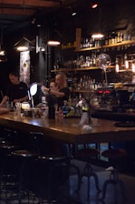 A dimly lit bar scene with two bartenders preparing drinks behind a wooden counter. The bar is stocked with various bottles on shelves, and the lighting creates a cozy atmosphere. Several hanging pendant lights illuminate the area, and there are bar stools lined up in front of the counter.