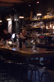 A dimly lit bar scene with two bartenders preparing drinks behind a wooden counter. The bar is stocked with various bottles on shelves, and the lighting creates a cozy atmosphere. Several hanging pendant lights illuminate the area, and there are bar stools lined up in front of the counter.