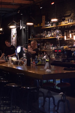 A dimly lit bar scene with two bartenders preparing drinks behind a wooden counter. The bar is stocked with various bottles on shelves, and the lighting creates a cozy atmosphere. Several hanging pendant lights illuminate the area, and there are bar stools lined up in front of the counter.