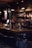 A dimly lit bar scene with two bartenders preparing drinks behind a wooden counter. The bar is stocked with various bottles on shelves, and the lighting creates a cozy atmosphere. Several hanging pendant lights illuminate the area, and there are bar stools lined up in front of the counter.