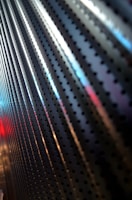 A close-up of a metallic surface with a series of round perforations, creating a repetitive pattern. The surface reflects light with shades of blue, red, and white creating a dynamic visual effect.
