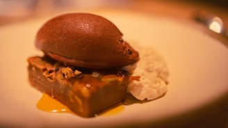 A rich dessert featuring a dark chocolate ice cream scoop placed on a rectangular piece of cake with nuts and caramel. Accompanied by a dollop of cream, the dessert is artfully arranged on a white plate with warm lighting.