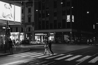 A nighttime urban scene with a black and white aesthetic, featuring a pedestrian crossing with a couple walking across. Neon signs illuminate the storefronts in the background, along with bicycles and a few people. An illuminated billboard displays an advertisement.