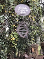 An ornate metal archway decorated with intricate wrought iron scrollwork stands amidst lush greenery. A round sign at the top reads 'Caminho das Abelhas' in elegant script. Hanging below the sign is a metal sculpture resembling a beehive with two bees. Vines with green and yellow leaves climb over the structure, and a stone pathway can be seen behind.