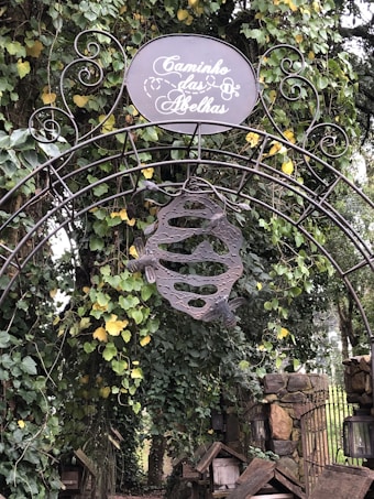An ornate metal archway decorated with intricate wrought iron scrollwork stands amidst lush greenery. A round sign at the top reads 'Caminho das Abelhas' in elegant script. Hanging below the sign is a metal sculpture resembling a beehive with two bees. Vines with green and yellow leaves climb over the structure, and a stone pathway can be seen behind.