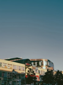 A modern building with large windows reflecting the sky, surrounded by other commercial structures with signs and advertisements. A few trees are visible in the foreground, casting shadows on the building. The sky is clear and blue, suggesting a sunny day.