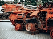 A rusted, heavily damaged military vehicle is shown with its internal structures exposed. The vehicle sits on a paved surface with another similarly wrecked vehicle in the background. The scene appears to be an outdoor area surrounded by trees.