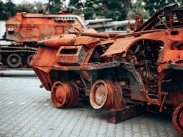 A rusted, heavily damaged military vehicle is shown with its internal structures exposed. The vehicle sits on a paved surface with another similarly wrecked vehicle in the background. The scene appears to be an outdoor area surrounded by trees.