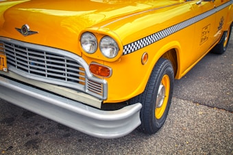 A vintage yellow taxi with a distinct checkerboard stripe runs along its side. The front section is prominently featured with a classic grille and dual round headlights. The chrome and bright paint give it a polished and nostalgic look.
