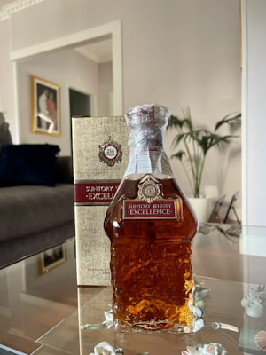 A bottle of Suntory Whisky Excellence is prominently displayed on a glass surface, with its label clearly visible. The bottle is paired with its packaging box, both of which feature an elegant design. In the background, a comfortable living room setting is visible, including a framed picture on the wall, a couch with dark cushions, and a potted plant.