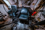 The interior of a tank or armored vehicle is visible, with two black leather seats positioned in a small, enclosed metal space. Various levers, controls, and mechanical parts are scattered around the floor and sides. A fire extinguisher is mounted to the right side, and wires and cables run along the walls.