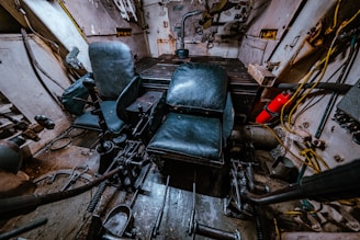 The interior of a tank or armored vehicle is visible, with two black leather seats positioned in a small, enclosed metal space. Various levers, controls, and mechanical parts are scattered around the floor and sides. A fire extinguisher is mounted to the right side, and wires and cables run along the walls.