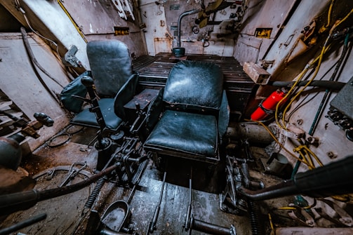 The interior of a tank or armored vehicle is visible, with two black leather seats positioned in a small, enclosed metal space. Various levers, controls, and mechanical parts are scattered around the floor and sides. A fire extinguisher is mounted to the right side, and wires and cables run along the walls.