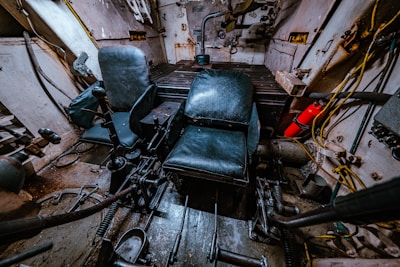 The interior of a tank or armored vehicle is visible, with two black leather seats positioned in a small, enclosed metal space. Various levers, controls, and mechanical parts are scattered around the floor and sides. A fire extinguisher is mounted to the right side, and wires and cables run along the walls.