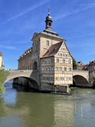 A historic building with intricate timber-framed architecture stands on a bridge over a river. The structure has a steep gabled roof and is adorned with numerous windows. The setting suggests a quaint, traditional town atmosphere, with clear, blue skies overhead.