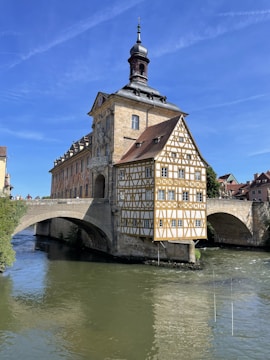 A historic building with intricate timber-framed architecture stands on a bridge over a river. The structure has a steep gabled roof and is adorned with numerous windows. The setting suggests a quaint, traditional town atmosphere, with clear, blue skies overhead.