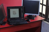 A workspace featuring a black laptop displaying a logo design, positioned on a red desk beside a black mouse and two additional monitors, one turned off and one angled towards a window with a grid pattern.