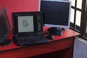 A workspace featuring a black laptop displaying a logo design, positioned on a red desk beside a black mouse and two additional monitors, one turned off and one angled towards a window with a grid pattern.