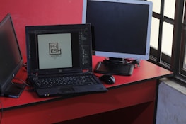 A workspace featuring a black laptop displaying a logo design, positioned on a red desk beside a black mouse and two additional monitors, one turned off and one angled towards a window with a grid pattern.