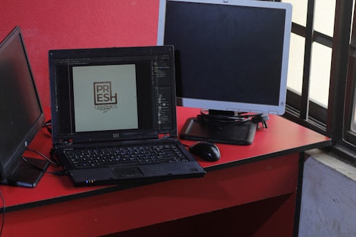 A workspace featuring a black laptop displaying a logo design, positioned on a red desk beside a black mouse and two additional monitors, one turned off and one angled towards a window with a grid pattern.