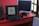A workspace featuring a black laptop displaying a logo design, positioned on a red desk beside a black mouse and two additional monitors, one turned off and one angled towards a window with a grid pattern.