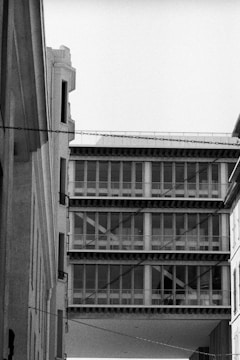 A black and white photograph capturing modern architecture with a prominent building featuring large windows and vertical lines. The structure appears to be a bridge between two adjacent buildings, creating a suspended corridor. Details such as balconies and architectural patterns are visible.