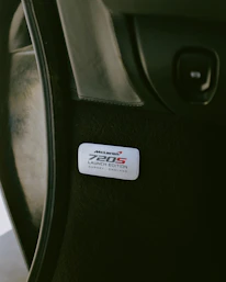 An interior vehicle badge with the text 'McLaren 720S Launch Edition Surrey, England' displayed on a dark surface. The badge is sleek and metallic, suggesting a premium, high-performance vehicle.