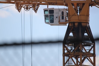 A close-up of a tower crane's cabin and structure against a blue sky backdrop, with some cables visible.