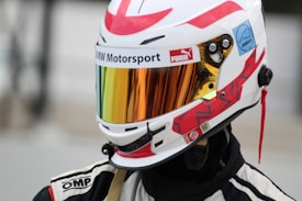 A person is wearing a white racing helmet with red accents and a reflective visor. The helmet features various logos and branding, including BMW Motorsport, Puma, and other designs. The person is also wearing a black and white racing suit with visible shoulder patches.