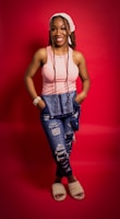 A person stands against a vibrant red backdrop, wearing a pink sleeveless top, distressed blue jeans, and beige fuzzy slippers. They have long braided hair and a light bandana, with hands tucked into their pockets. The person's expression is cheerful and relaxed.