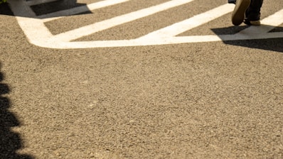 A section of an asphalt pavement featuring bold white traffic markings. A pair of shoes is partially visible in the top right, suggesting someone walking across the marked area.