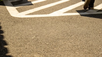 A section of an asphalt pavement featuring bold white traffic markings. A pair of shoes is partially visible in the top right, suggesting someone walking across the marked area.