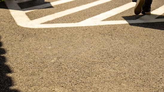 A section of an asphalt pavement featuring bold white traffic markings. A pair of shoes is partially visible in the top right, suggesting someone walking across the marked area.