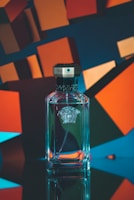 A clear glass perfume bottle with a black octagonal cap is placed against a backdrop of abstract geometric shapes in vibrant colors, predominantly blue and orange. The bottle has an embossed logo on the front, and the lighting creates a reflective effect on the surface.