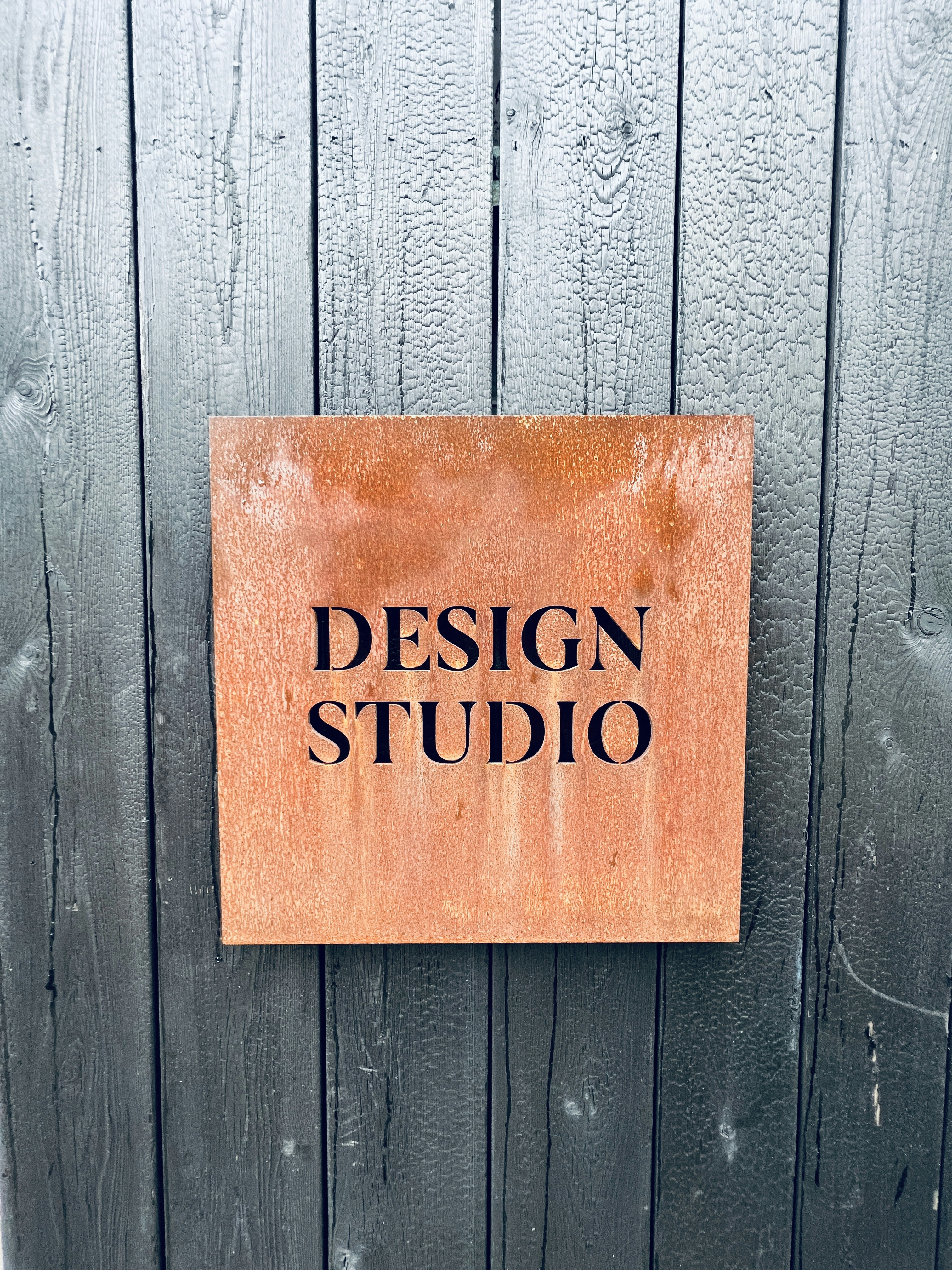 Rostig metallskylt för designstudio monterad på ett sotigt trästaket.
