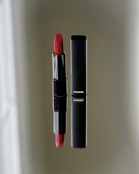 A vibrant red lipstick stands next to its sleek black case on a reflective surface. The brand name is visible on the case, emphasizing luxury and elegance.