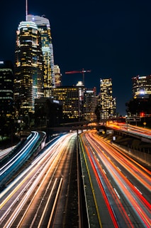 A vibrant urban nightscape featuring tall skyscrapers illuminated against a dark sky. In the foreground, long exposure captures light trails from vehicles on a busy highway, creating dynamic streaks of red and white. The city is alive with energy, marked by numerous buildings glowing with office lights.