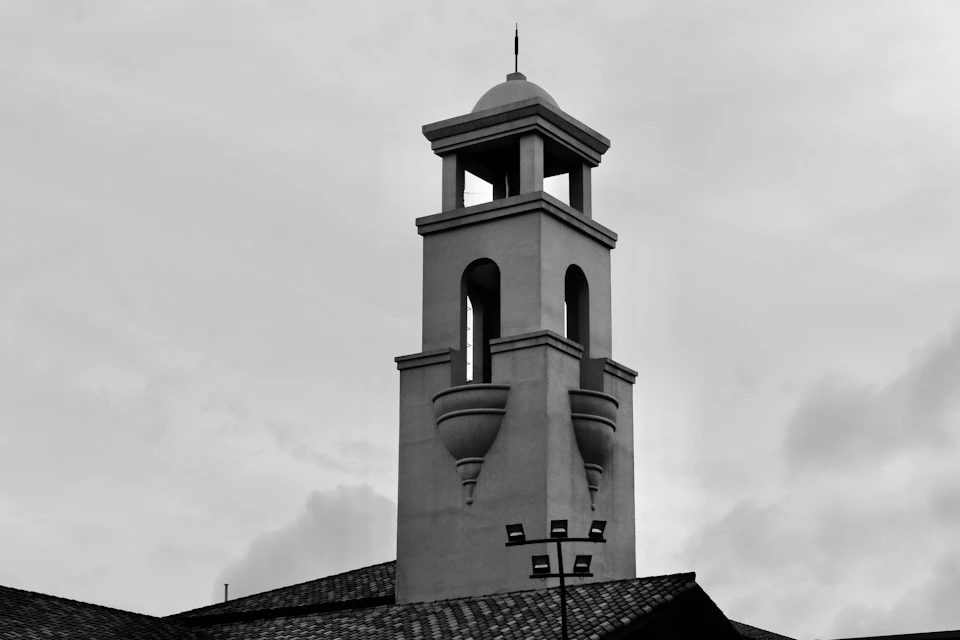 The image depicts a tall, white bell tower with a pointed roof and two windows on each side. The tower is situated atop a building with a tiled roof, adding to its architectural charm. The photo is taken from below, emphasizing the height of the tower against an overcast sky.