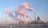 Several tall smokestacks emit thick plumes of smoke into the sky, with a backdrop of an urban area. A red building with a cross on top stands to the right, resembling a church. The sky is mostly clear with a hint of pollution.