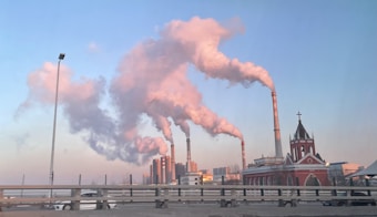 Several tall smokestacks emit thick plumes of smoke into the sky, with a backdrop of an urban area. A red building with a cross on top stands to the right, resembling a church. The sky is mostly clear with a hint of pollution.
