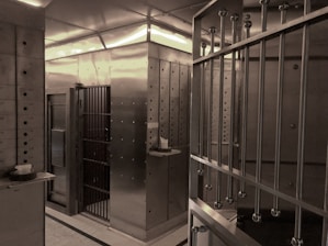 An interior space dominated by metal surfaces and structures, resembling a vault or secure facility. The walls and door are made of metal, with visible rivets and steel bars forming a secure area. There are numbered boxes on the left, possibly indicating storage or safety deposit boxes, and a shelf with a small object or item placed on it.